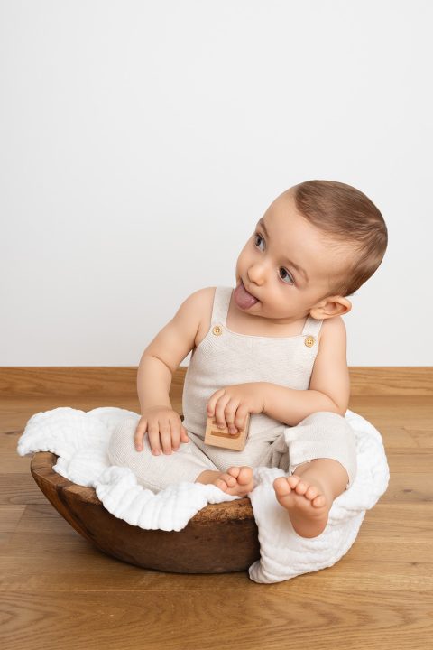 Ein einjähriges Baby sitzt in einer Holzschale und zeigt die Zunge.